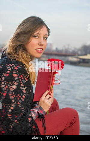 Belle fille brune avec un livre près de la rive du fleuve holding red flower Banque D'Images