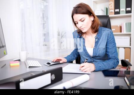 Femme travailleuse assise à son bureau dans le bureau de rédiger un rapport et de faire des calculs sur une calculatrice manuelle Banque D'Images