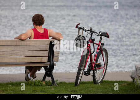 Vue de l'arrière d'un homme en rouge T-shirt de muscle assis sur un banc, se reposant après la randonnée à vélo. Banque D'Images