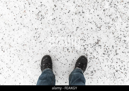 Les pieds mâles en jean bleu et des chaussures noires debout sur la route d'hiver enneigé avec lame de gravillons de granit, vue à la première personne Banque D'Images