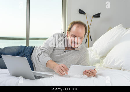 Man Lying in Bed smiling travaillant sur ordinateur portable et tablette Banque D'Images