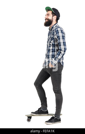 Vue latérale du hipster barbu avec cap debout sur la planche à roulettes à la voiture en souriant. La pleine longueur du corps portrait isolated over white Banque D'Images