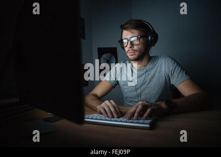Jeune homme concentré dans les écouteurs assis dans une pièce sombre et à l'aide d'ordinateur Banque D'Images