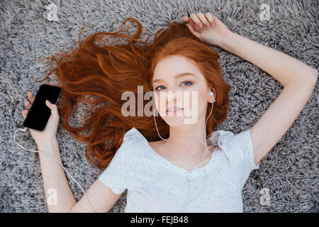 Belle femme rousse allongée sur le tapis et l'écoute de la musique sur le smartphone à la maison Banque D'Images