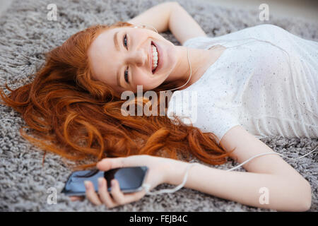 Smiling redhead woman allongé sur le tapis et l'écoute de la musique sur le smartphone à la maison Banque D'Images