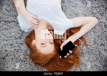 Belle femme rousse allongée sur le tapis et l'écoute de la musique sur le smartphone. Banque D'Images