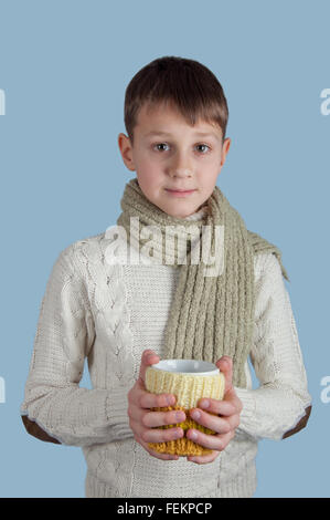 Cute boy en blanc pull et écharpe verte avec une tasse blanche sur fond bleu Banque D'Images