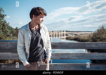 Young man leaning against clôture en bois sur le pont sur la rivière, les mains dans les poches à la recherche de là, Costa Smeralda, Sardaigne, Italie Banque D'Images