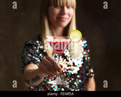 Mid adult woman holding jus cru dans verre à cocktail garni d'une tranche de lime smiling at camera Banque D'Images