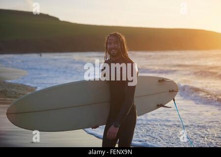 Portrait of young male surfer carrying surfboard on beach, Devon, England, UK Banque D'Images