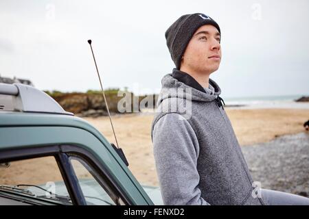 Young man leaning against car le regard vers le haut de la plage, à Constantine Bay, Cornwall, UK Banque D'Images