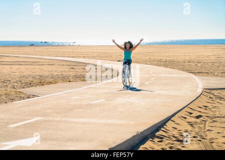 Mid adult woman en vélo le long de sentiers, les bras dans l'air de la plage Banque D'Images