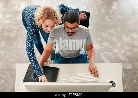 Vue de dessus coup de jeune homme assis à son bureau travaillant sur pc et tablette graphique numérique avec femme pointant sur moniteur. Homme gr Banque D'Images