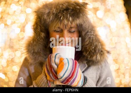 Young woman wearing furry hood et mitaines holding boisson chaude en face de lumières de Noël Banque D'Images