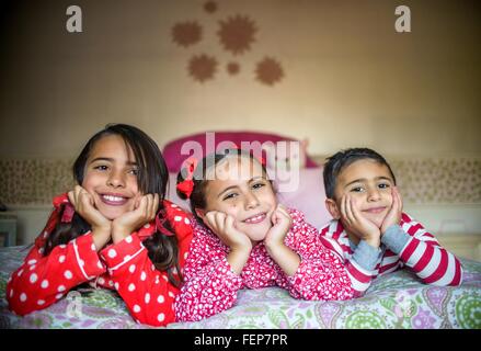 Enfants vêtus de pyjamas couché sur fronts sur menton lit dans les mains à la caméra en souriant Banque D'Images