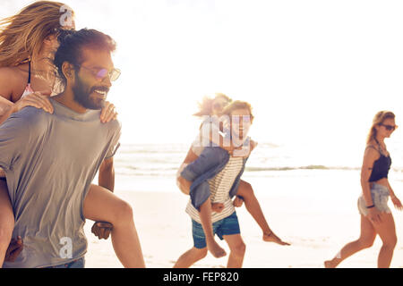 Groupe d'amis s'amuser sur la plage, les jeunes hommes sur le dos des femmes sur le bord de la mer. Mixed Race jeunes bénéficiant de l'été Banque D'Images