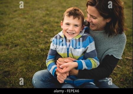 Garçon assis sur les genoux des mères sur l'herbe à la caméra en souriant Banque D'Images