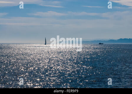 Voilier et bateau à moteur sur l'eau reflétant ondulée contre rive brumeuse lointain et partiellement nuageux ciel. Banque D'Images