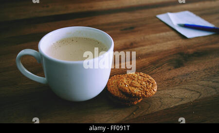 Un latte mousseux est assis à côté d'une assiette de biscuits, disposés dans une composition de nature morte. La tasse blanche contient la boisson crémeuse, créant une scène chaleureuse et accueillante. L'accent est mis sur la boisson chaude et réconfortante associée aux biscuits sucrés. Banque D'Images