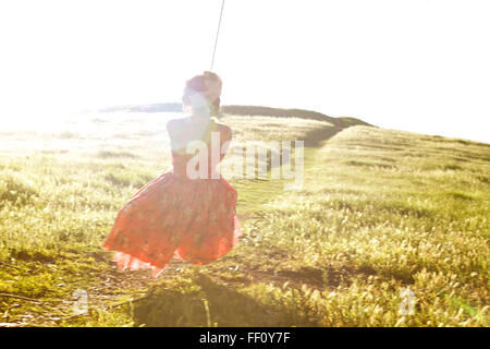 Vue de derrière d'une jeune fille dans une robe rose à fleurs se balançant sur une corde balançoire avec un chemin à travers un terrain ensoleillé à l'arrière-plan. Banque D'Images