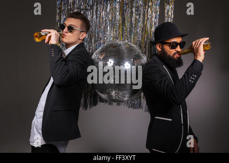 Deux beaux jeunes hommes confiants en costumes noirs et lunettes, debout près de boule disco et boire de la bière sur fond noir Banque D'Images