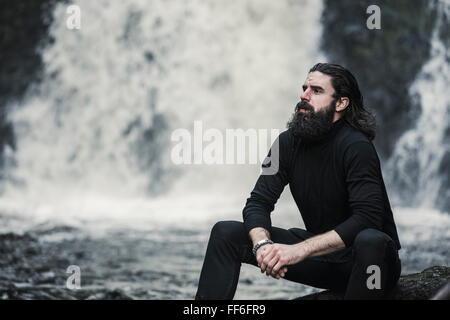 Un homme assis avec ses mains jusqu'à la recherche, par une cascade d'eau, un ruisseau de montagne. Banque D'Images