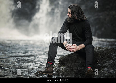 Un homme assis par un jet d'eau qui coule sur les rochers. Banque D'Images