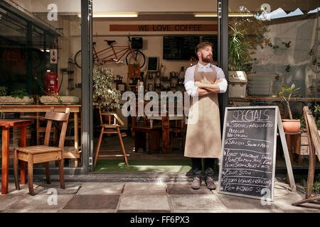 Portrait d'un jeune homme debout devant son café. Jeune homme avec barbe portant un tablier, debout avec ses bras cros Banque D'Images