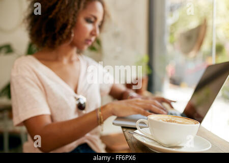 Tasse de cappuccino sur table avec une jeune femme travaillant sur ordinateur portable en arrière-plan. L'accent de tasse de café au café. Banque D'Images