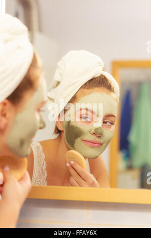 Woman argile séchée du visage masque de boue avec éponge dans la salle de bains en face de miroir. Soins de la peau. Fille prendre soin de son visage. Banque D'Images