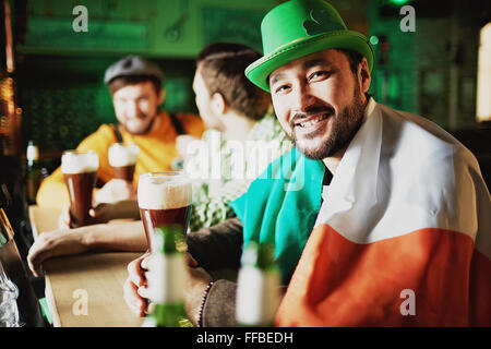 Young Asian man avec verre de bière et drapeau irlandais assis dans un pub Banque D'Images