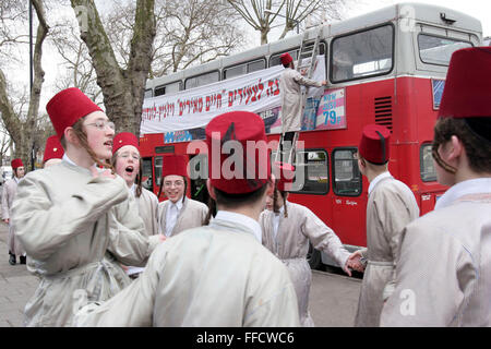 Un groupe de garçons juifs orthodoxes de la Yeshiva (école) Viznitz habillé en robe de chanter et danser dans la rue. D'autres membres du groupe décorent les bus à deux étages avec des bannières qu'ils voyageront sur pour la fête juive de Pourim. Ils visiteront plusieurs riches hommes d'affaires locaux à recueillir des fonds pour leur école. Banque D'Images