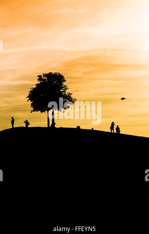 Silhouettes de personnes sur un monticule, arbre et un enfant flying a kite au crépuscule Banque D'Images