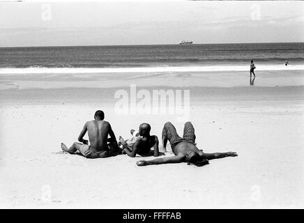 Jan 4, 2016 - South Beach, KwaZulu-Natal, Afrique du Sud - trois jeunes hommes se baigner de soleil avec un bateau et un homme marchant dans l'arrière-plan. South Beach est une partie de la ville de Durban sans interruption la plus longue étendue de plage de sable. La ville de Durban est sur la côte est de l'Afrique du Sud et les gens ici sont lavés avec de l'eaux chaudes de l'Océan Indien. Au nord de cette étendue de sable sont des plages avec la société hang out cafe. Au sud il y a une jetée avec le quartier haut Moyo's Restaurant à elle est la fin et l'uShaka Marine World et le secteur complexe de la mer et surf clubs de la vesces Banque D'Images