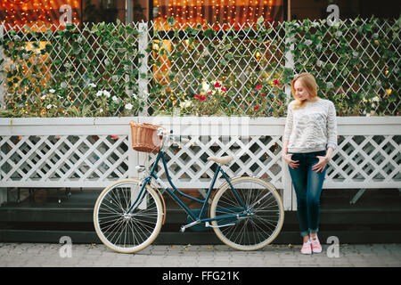 Jeune femme sérieuse sur un vélo à l'époque Ville Rue debout près de Café de la rue avec mur de fleurs. Focus sélectif. Banque D'Images