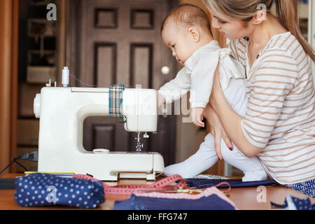 Petit enfant apprend de nouvelles connaissances, de même que sa mère inspecte la machine à coudre. Travailler à la maison, éducation des enfants, les parents et les enfants, de soins, de baby-sitter. Banque D'Images