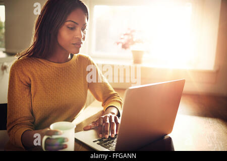 Noir femme célibataire sérieux adultes assis à table holding Coffee cup et de la saisie sur ordinateur portable avec flare lumière venant par windo Banque D'Images