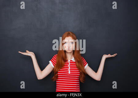 Assez confus jeune femme avec des longs cheveux rouges holding copyspace sur les deux paumes sur arrière-plan du tableau Banque D'Images