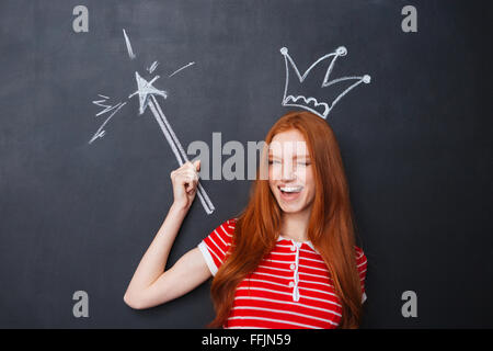 Jolie jeune femme mignonne et clignant debout avec couronne et baguette magique sur l'arrière-plan du tableau Banque D'Images