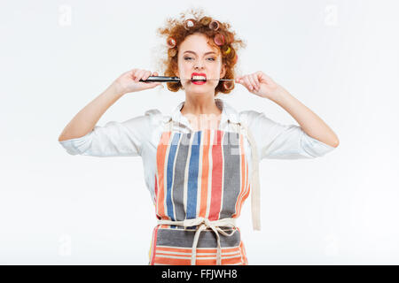 Belle jeune femme au foyer de curly holding tablier de cuisine et de mordre grand couteau sur fond blanc Banque D'Images