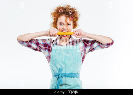 Jolie rousse femme au foyer en chemise à carreaux et tablier bleu holding et mordants sur fond blanc spaghettis non cuits Banque D'Images