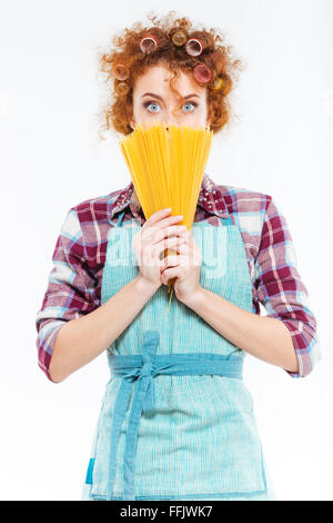 Portrait de jeune femme au foyer assez étonné avec bigoudis en tablier bleu couvert le visage avec spaghettis non cuits sur fond blanc Banque D'Images