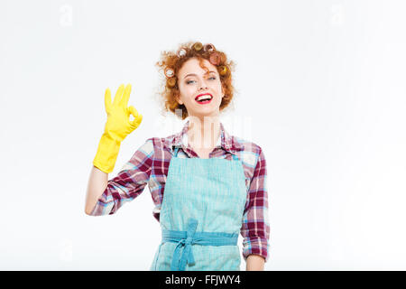 Jeune femme au foyer heureux curly en tablier bleu et des gants jaunes montrant ok sur fond blanc Banque D'Images