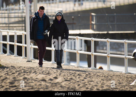 Aberystwyth, Pays de Galles, Royaume-Uni. 15 février 2016. Météo France : deux personnes de profiter d'une journée d'hiver au chaud soleil de marcher le long de la promenade à Aberystwyth, qui est encore partiellement recouverte de sable et de galets lavés par la tempête féroce Imogen il y a exactement une semaine. De nombreuses écoles ont pris leurs vacances cette semaine. Credit : Keith morris/Alamy Live News Banque D'Images