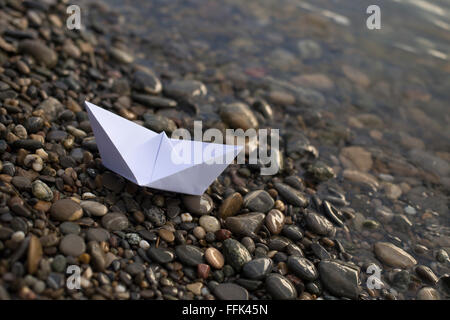 Un petit bateau de papier blanc sur les cailloux sur la rive d'une rivière Banque D'Images