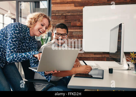 Photo de deux jeunes heureux graphistes travaillant dans leurs bureaux à l'homme assis à son bureau et collègue montrant certains Banque D'Images