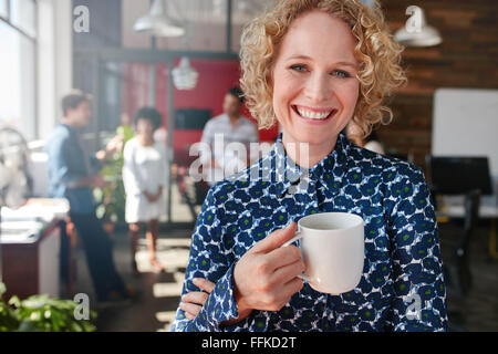 Portrait of smiling female designer tenant une tasse de café. Elle se tient à l'office de tourisme avec des collègues discutant en arrière-plan Banque D'Images