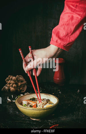 En main de femme chemise rouge prendre par baguettes rouge de nouilles ramen Soupe Asiatique aux crevettes, l'oignon, des tranches de champignons et d'œufs, la serv Banque D'Images