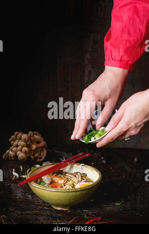 En main de femme chemise rouge parsemer de tranches de ciboule soupe ramen Asiatique aux crevettes, nouilles, des tranches de champignons et d'œufs, la serv Banque D'Images