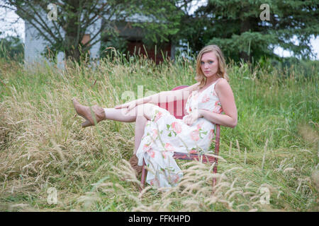 Belle blonde femme assis sur un vieux fauteuil rouge dans les champs. Banque D'Images